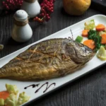 "A whole grilled dorado fish on a white rectangular plate, accompanied by colorful vegetables"