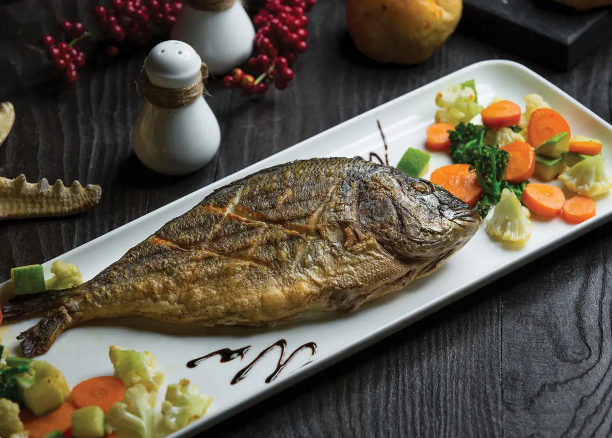 "A whole grilled dorado fish on a white rectangular plate, accompanied by colorful vegetables"