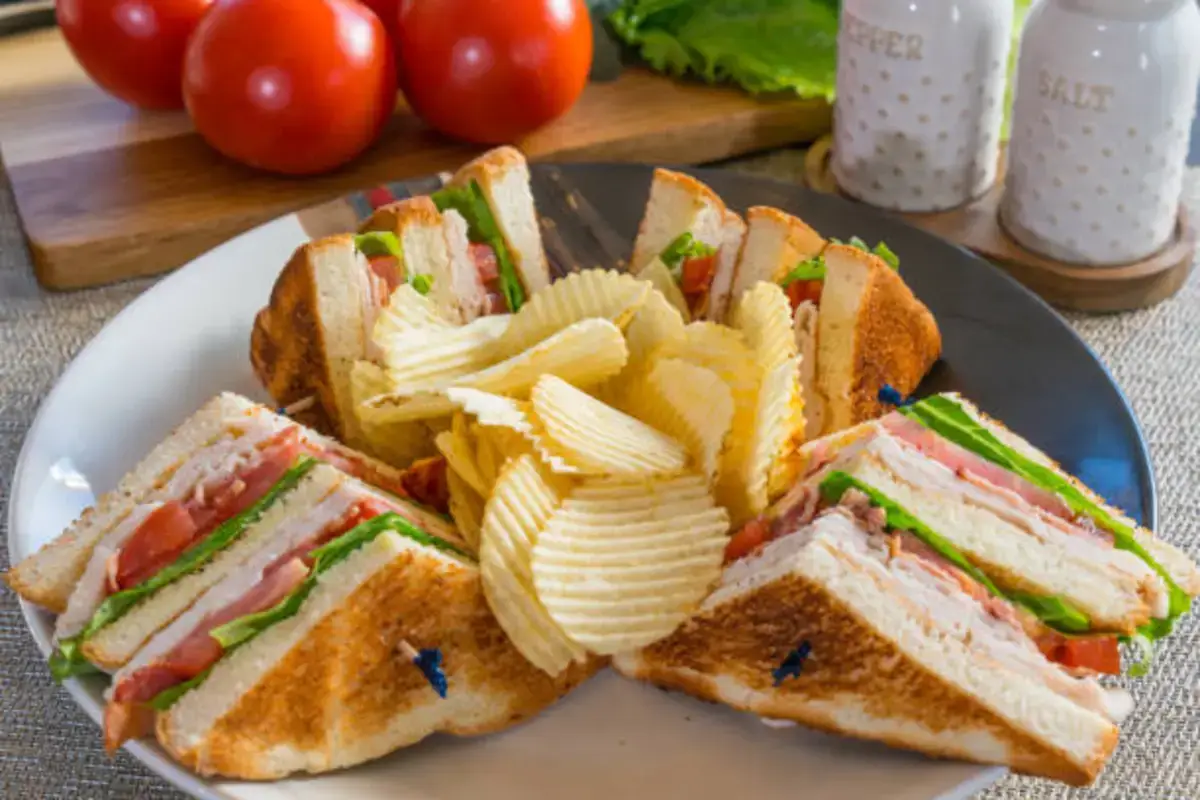 "Club sandwich slices arranged around potato chips on a plate with fresh tomatoes in the background."